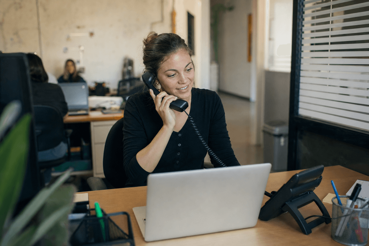 A small business sales rep picks up the phone to respond to a new lead notification while a laptop pipeline board sits open on the desk.